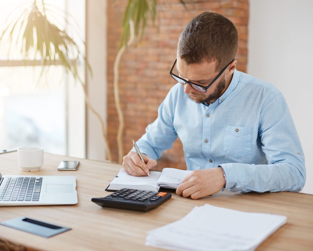 business concept. young unshaved caucasian male manager in glasses sitting in company office, writing down dates of meeting with customers in notebook with concentrated expression.